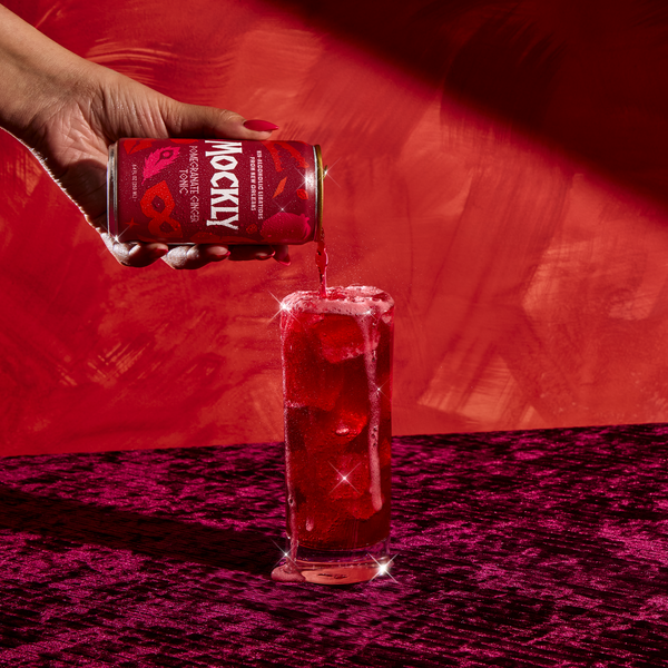 Hand holding a can of Pomegranate Ginger Tonic over a glass filled with red liquid on a red background