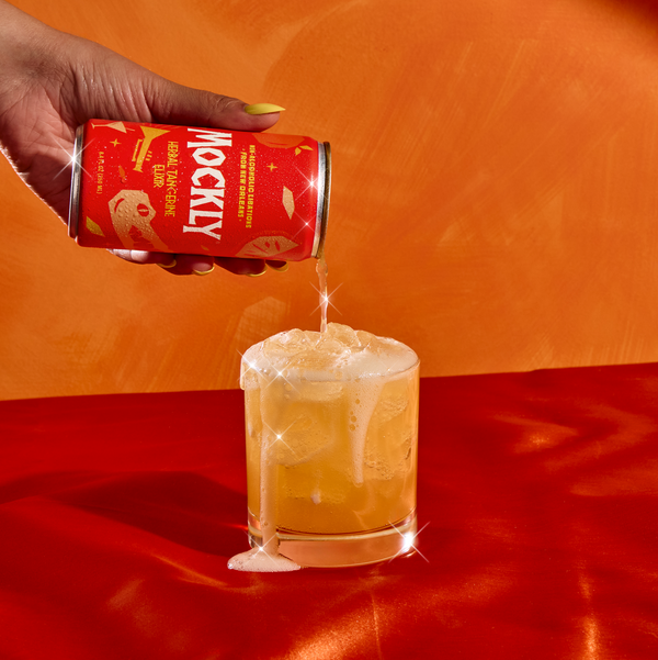 Hand pouring a drink from a can labeled Herbal Tangerine Elixir into a glass on an orange surface.